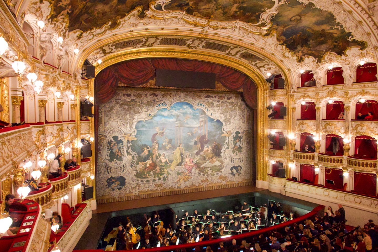 Ein Orchester spielt in einem verzierten, mehrstöckigen historischen Opernhaus.