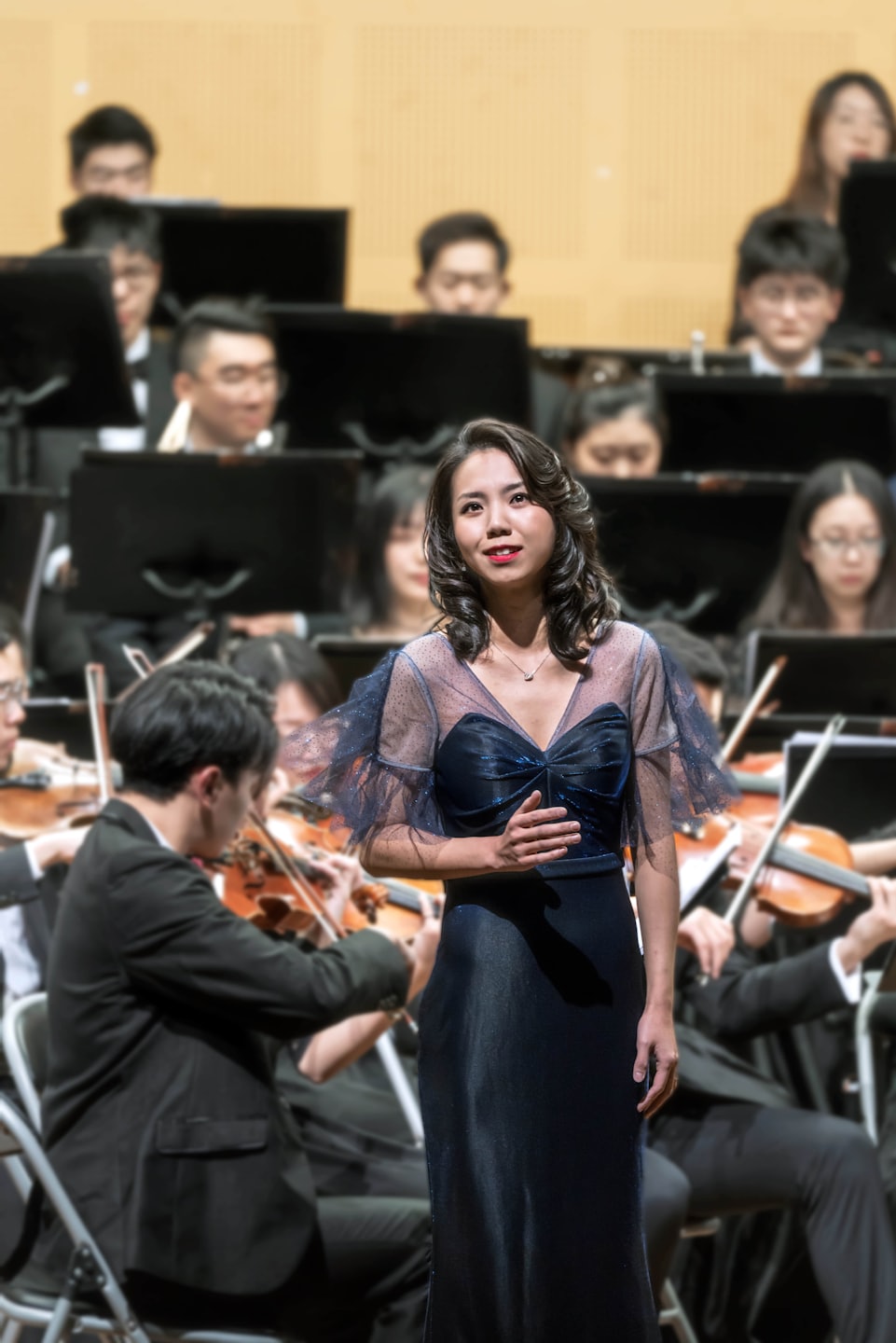 Eine Frau in einem blauen Kleid singt auf der Bühne mit einem unscharfen Orchester.